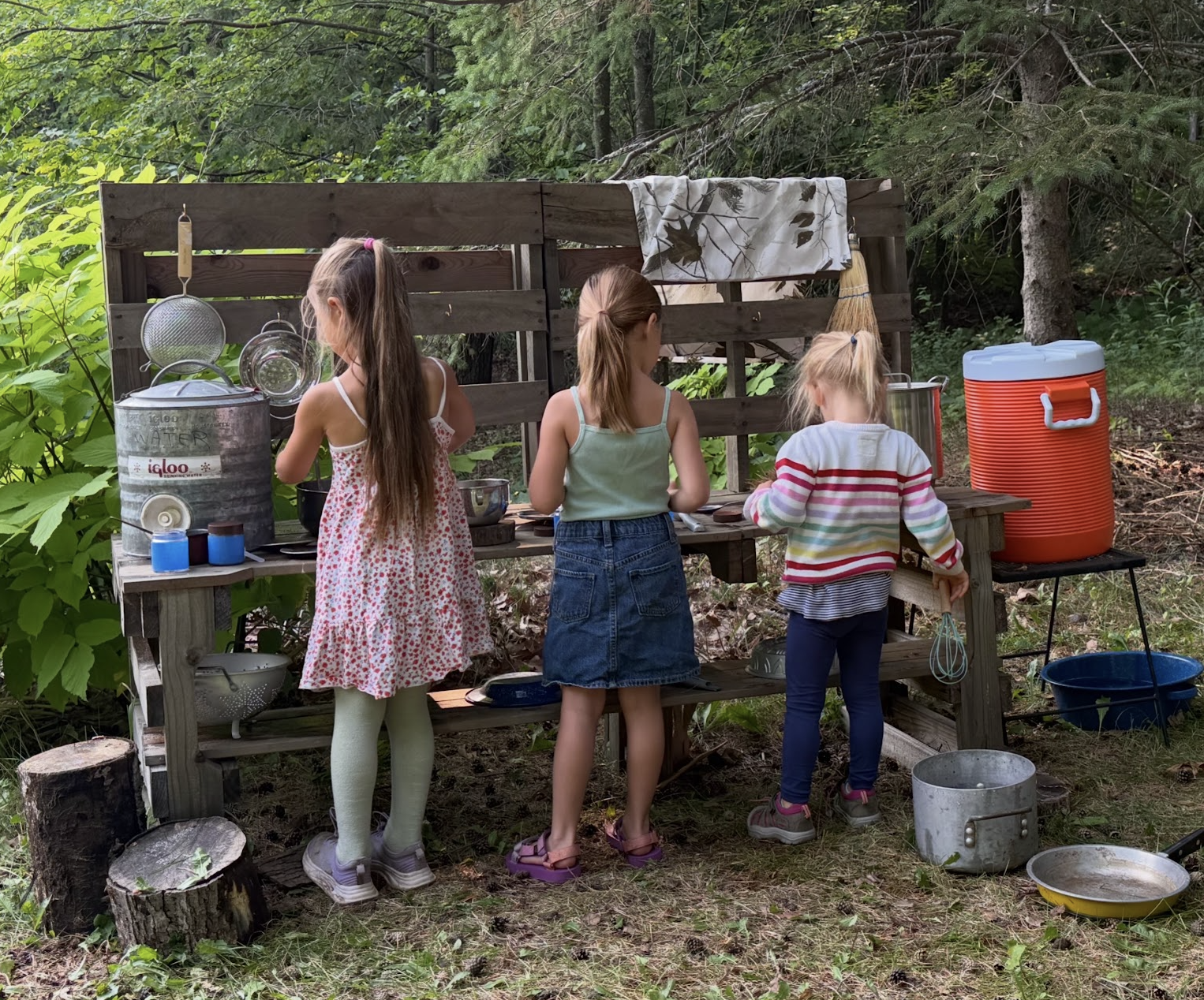 Three children playing at an outdoor mud kitchen surrounded by woods